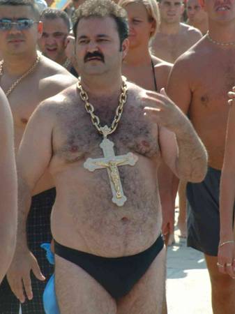 1986 Ford Bronco - A crowded beach scene showing a mustached man in a black speedo wearing an oversized gold chain with a very large crucifix pendant, standin…