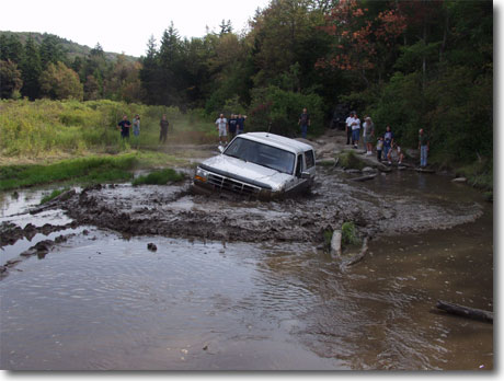 1995 Ford Bronco - A silver 1995 Ford Bronco navigates a muddy obstacle course, with onlookers observing from the wooded bank.