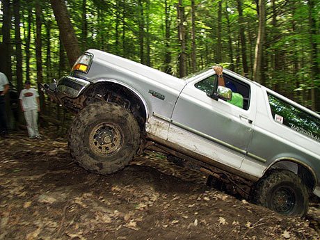 1995 Ford Bronco - A two-tone 1995 Ford Bronco navigates a challenging off-road trail through a dense forest.