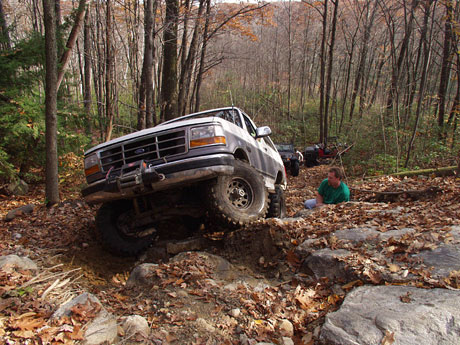 1995 Ford Bronco - A white Ford Bronco navigates a challenging off-road course in a forest, with another vehicle and a person visible in the background.