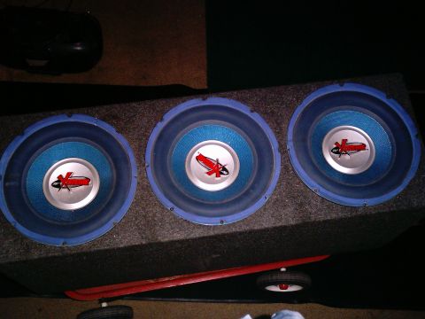 1986 Ford Bronco - Three blue-cone "Xtreme" car subwoofers with silver dust caps mounted in a gray carpeted three-hole enclosure, shot from directly above in…