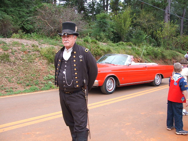 Man in a top hat and period suit stands next to a red convertible. A boy in a scout uniform is on the right