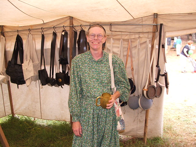 Woman in a green floral dress and bonnet holds a mug, standing in front of a tent displaying historical bags and clothing
