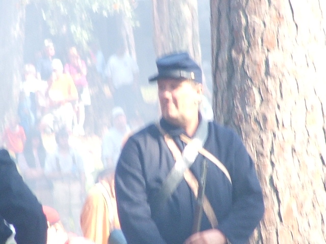 Man in a blue uniform and hat stands near a tree with a crowd in the background