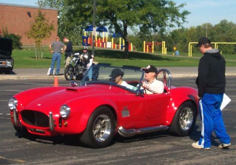 1979 Ford F-150 - A red convertible sports car with a roll bar is parked on asphalt with people around it.