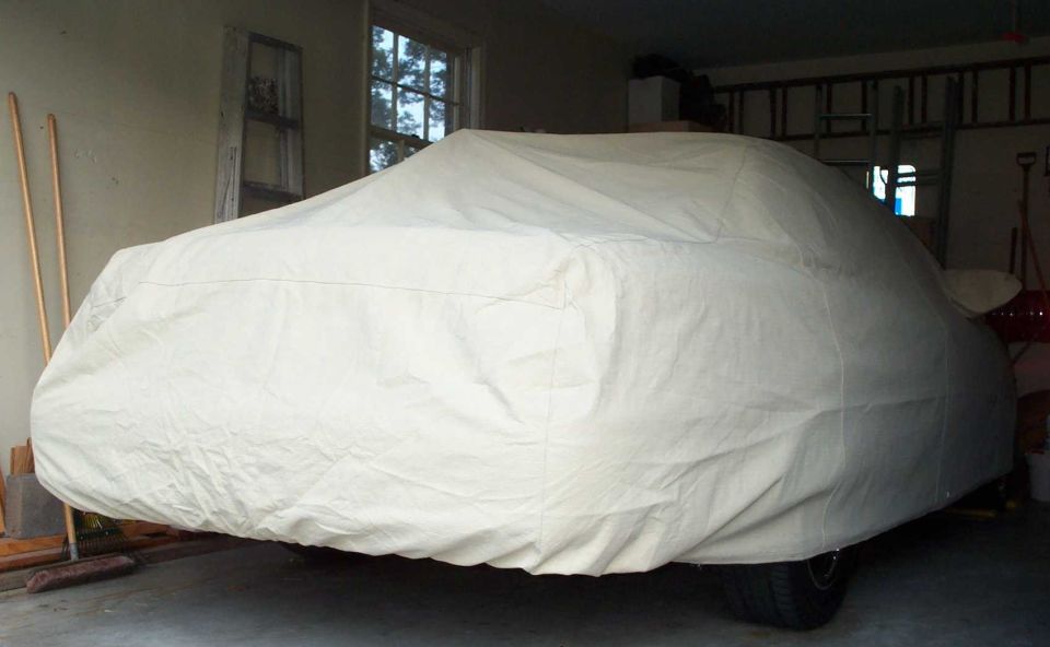 Car in a home garage fully hidden under a cream fabric car cover, one rear tire visible