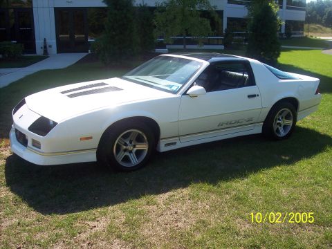 1988 Chevrolet Camaro - A white 1988 Chevrolet Camaro IROC-Z is shown from a three-quarter front view, parked on a grassy area in front of a modern building.