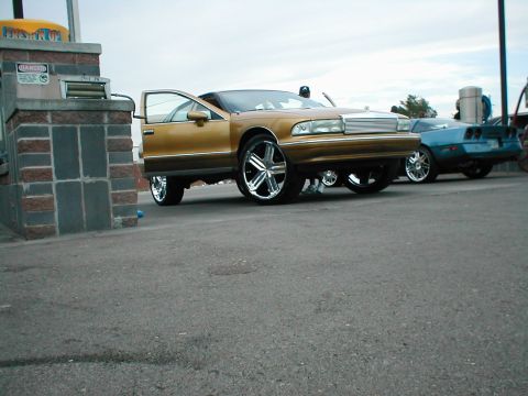 1979 Ford Bronco - A gold sedan with large chrome wheels is parked with its doors open, next to a blue sports car.