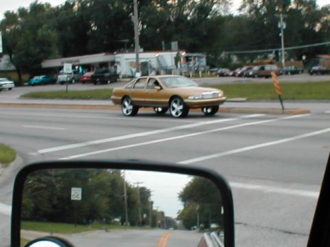 1979 Ford Bronco - Gold sedan with custom wheels drives down a street, as seen in a side mirror.