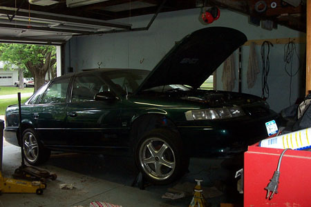 1995 Ford Taurus - A dark green 1995 Ford Taurus is parked in a garage with its hood open and is supported by jack stands.