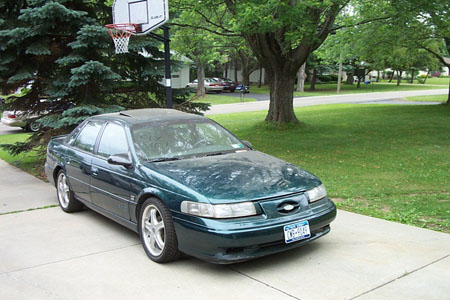 1995 Ford Taurus - Green 1995 Ford Taurus sits on a driveway next to a basketball hoop and trees.