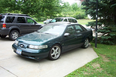 1995 Ford Taurus - A dark green sedan is parked on a concrete driveway, with an SUV to its left and a white vehicle behind it.