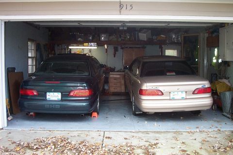 1995 Ford Taurus - Two 1995 Ford Tauruses are parked side-by-side in a garage, one dark green and the other tan.