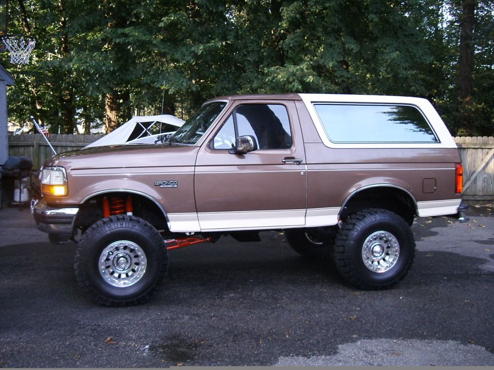 Tan Ford Bronco with white hardtop, lifted on mud tires, side profile by a fence