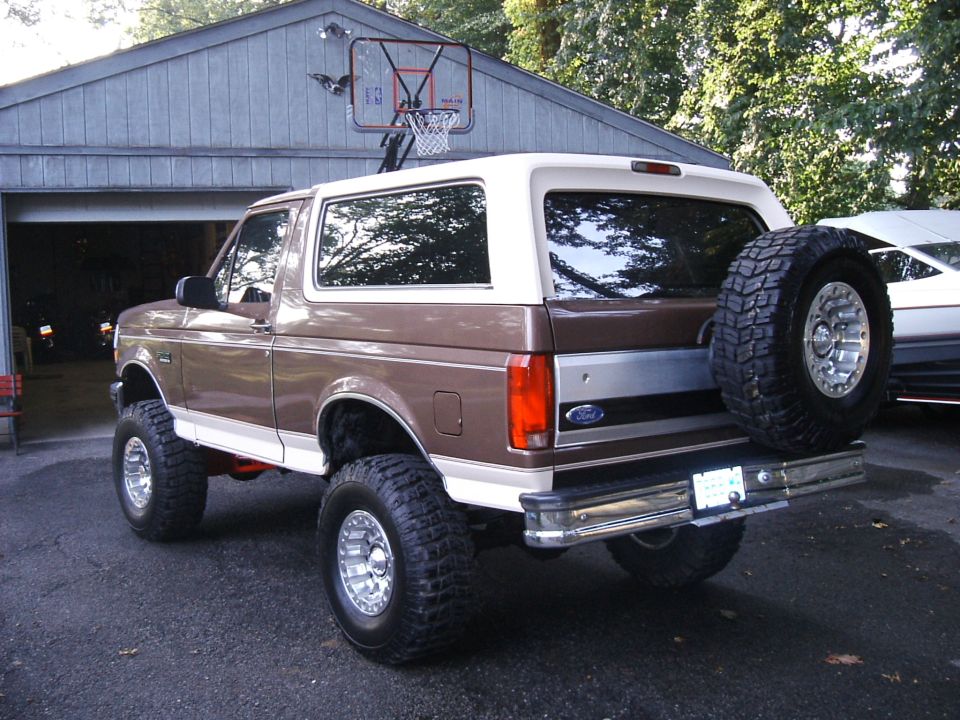 Lifted brown and tan Ford Bronco, rear three-quarter view, tailgate-mounted spare and mud tires, in a driveway