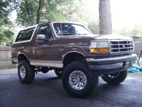 1993 Ford Bronco - This lifted Ford Bronco sits parked on a paved surface, with trees and a fence in the background.