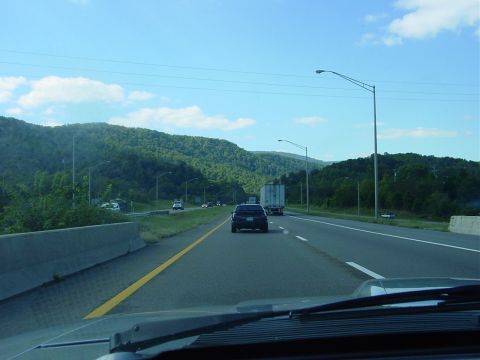 1986 Ford Mustang - View from a vehicle driving on a highway surrounded by forested mountains under a blue sky.