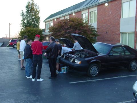 1986 Ford Mustang - A group of people gather around a dark red Ford Mustang with its hood open, possibly at a car meet or event.