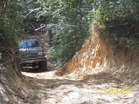 1987 Ford Bronco - A 1987 Ford Bronco navigates a narrow dirt path surrounded by trees and a steep embankment.