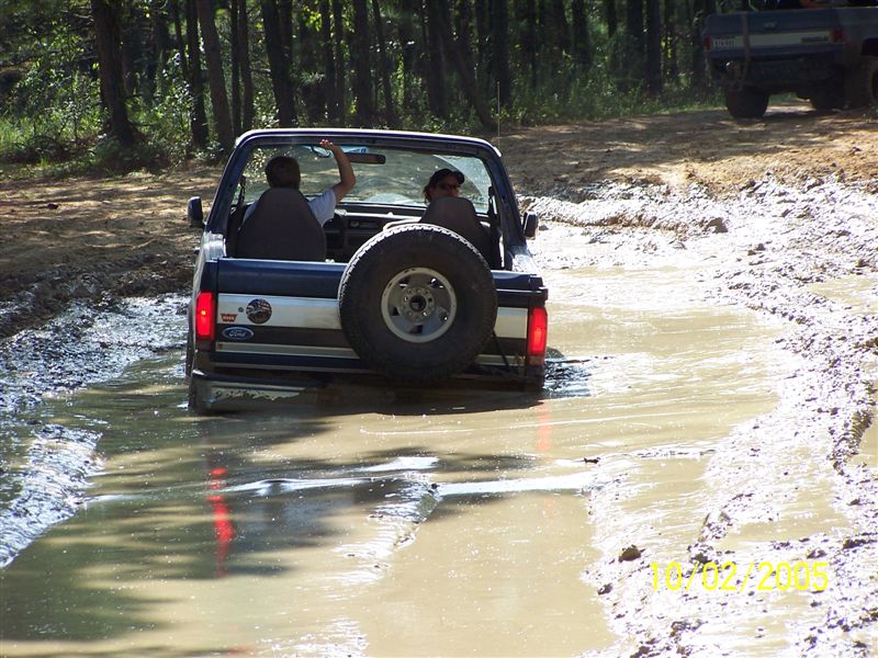 1987 Ford Bronco driving through a muddy trail with two people inside. Another truck is visible in the background