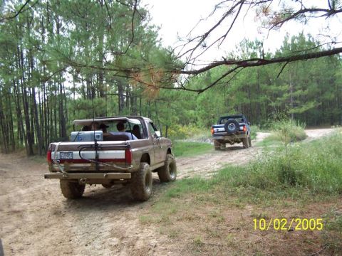 1987 Ford Bronco - Two trucks drive down a wooded dirt road on a cloudy day.