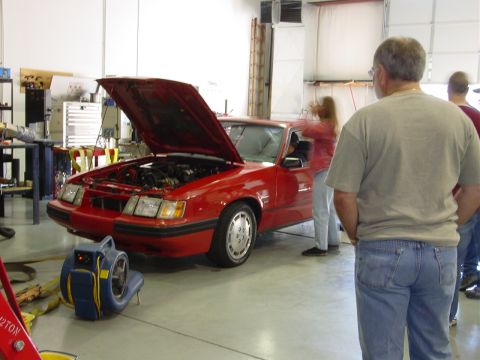 1986 Ford Mustang - A red 1986 Ford Mustang is displayed with its hood up, while onlookers observe.