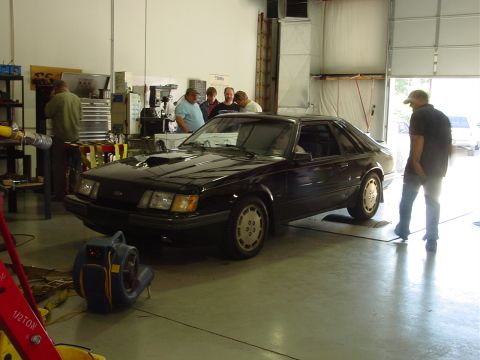 1986 Ford Mustang - A dark coupe with a hood scoop is parked in a garage, with several people observing.