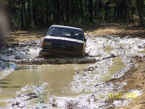 1987 Ford Bronco - A Ford truck navigates a muddy trail, kicking up water and dirt as it goes.