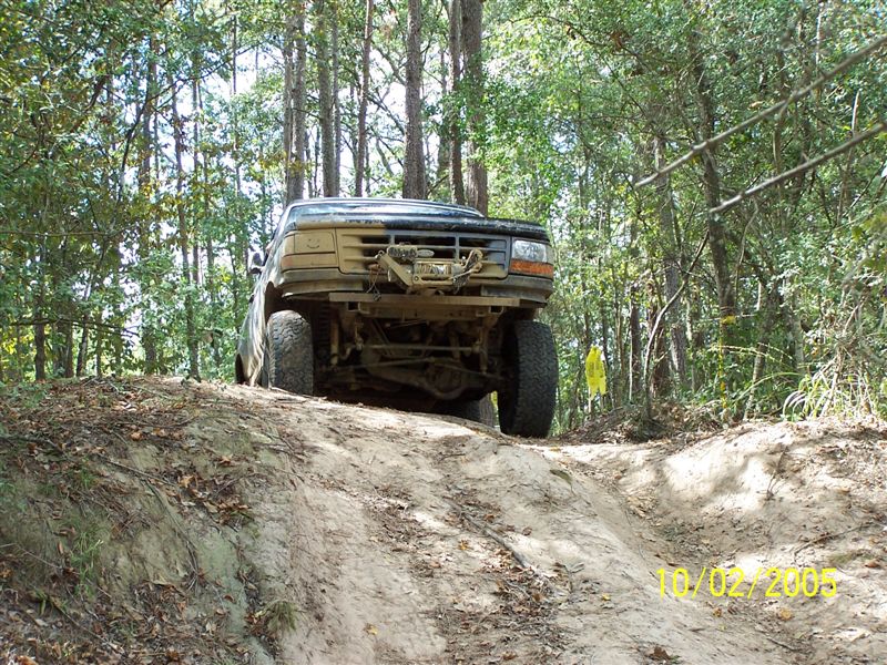 Front view of a lifted, mud-covered 1992 Ford Bronco driving on a dirt trail through woods. Winch is mounted on the front bumper