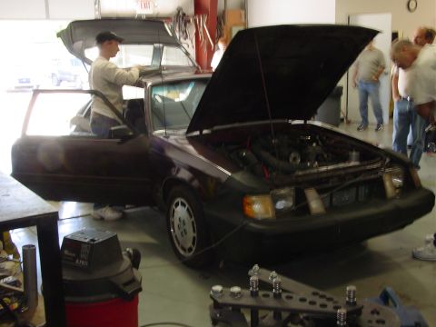 1986 Ford Mustang - A group of people examine the engine bay of a third-generation Ford Mustang in a garage.