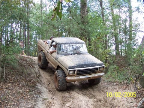 1987 Ford Bronco - A lifted Ford pickup truck navigates a muddy trail through a wooded area on a sunny day.