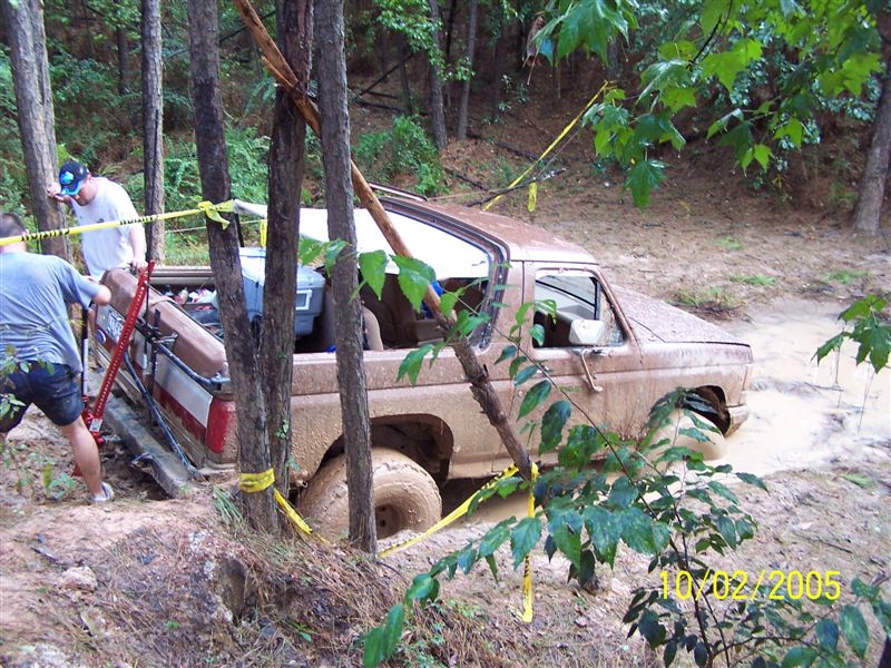 Mud-covered brown 1980s Ford Bronco stuck in a muddy ditch with two people nearby