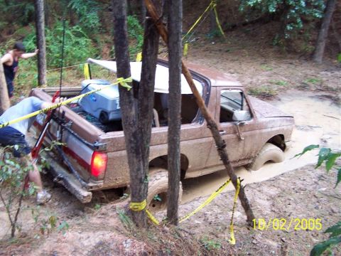 1987 Ford Bronco - A brown 1987 Ford Bronco is stuck deep in mud, with people attempting to help.