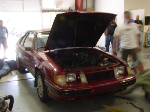 1986 Ford Mustang - A red hatchback, likely from the early 1980s, is parked with its hood open as people gather around.