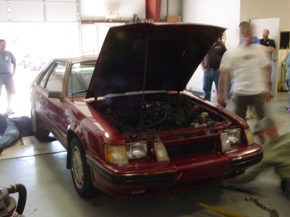 Red hatchback car, front view, hood open, showing engine. People around. Supermotors.net logo visible
