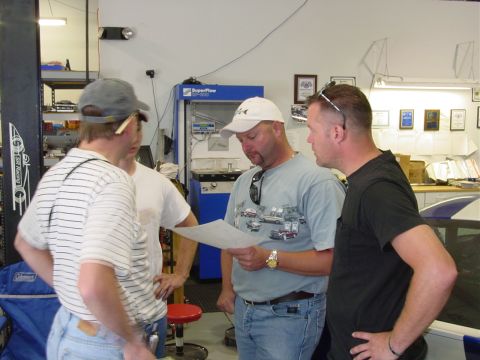 1986 Ford Mustang - Men examining a document in a workshop, with a car lift and equipment in the background.
