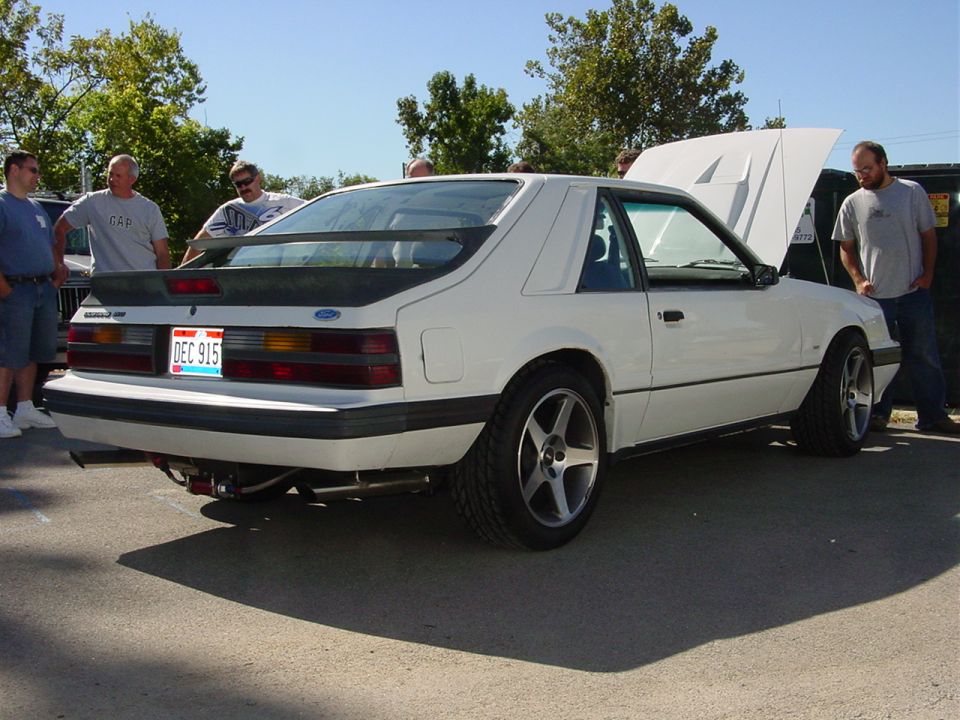 White 1987 Ford Mustang hatchback with a rear spoiler and aftermarket wheels, viewed from the rear three-quarter angle