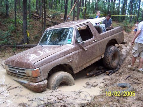 1987 Ford Bronco - Mud-caked brown Ford Bronco with its top removed and doors off, sunk into a rutted mud pit in a pine forest with logs wedged under the rear…