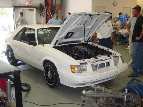 1986 Ford Mustang - A white 1986 Ford Mustang is displayed with its hood open, surrounded by onlookers in a garage setting.