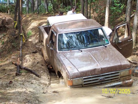 1987 Ford Bronco - A brown late-1980s Ford Bronco, mud-covered from bumper to roof and with its top removed, buried to the axles in a muddy rut in a pine wood…