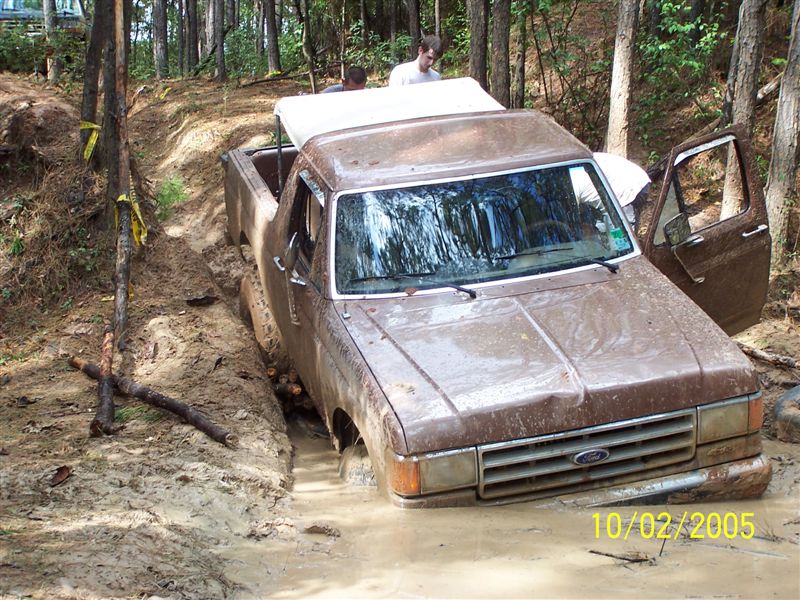 Mud-covered brown Ford Bronco stuck axle-deep in a muddy rut in the woods