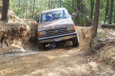 1987 Ford Bronco - This Ford truck is driving through a challenging off-road course with steep, muddy banks and water obstacles.