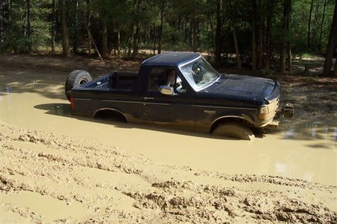 1987 Ford Bronco - A black 1987 Ford Bronco is stuck in a muddy area with trees in the background.