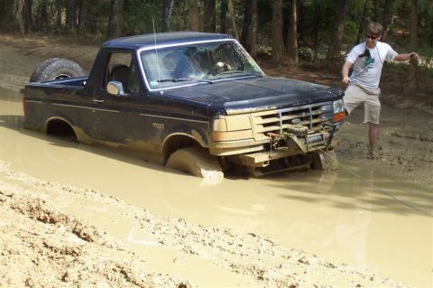 1987 Ford Bronco - A black 1987 Ford Bronco is stuck deep in the mud, with a person attempting to pull it out with a winch.