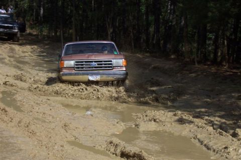 1987 Ford Bronco - A Ford truck navigates a challenging muddy path, with another vehicle following behind in the woods.