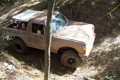 1987 Ford Bronco - A muddy Ford Bronco navigates an off-road trail with passengers visible inside.