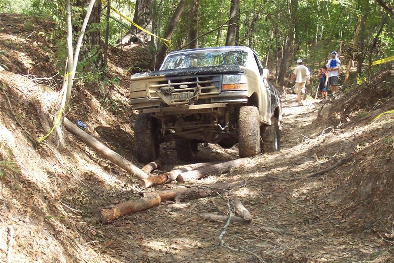 1987 Ford Bronco driving over logs on a dirt trail in woods