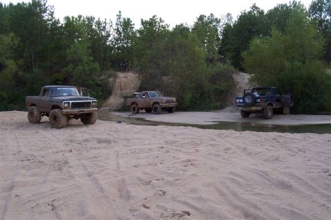 1987 Ford Bronco - Three lifted trucks are parked in a sandy area with trees and water, suggesting an off-road excursion.