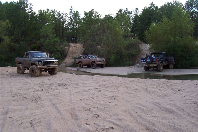 Three lifted trucks in a sandy area; front truck is a dark 1980s Ford F-Series