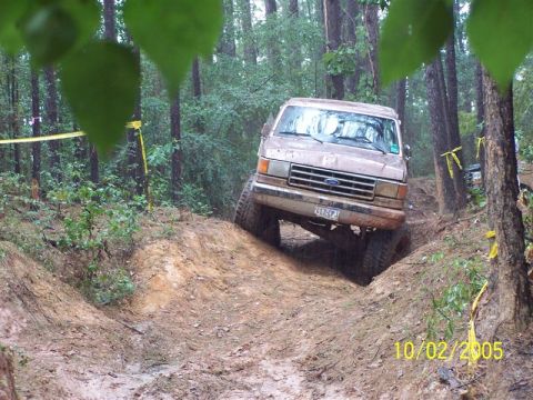 1987 Ford Bronco - A Ford Bronco navigates a challenging off-road trail on a wet day, with yellow caution tape visible in the background.